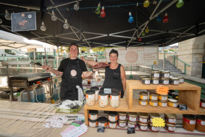 Deux productrices locales de miel posent devant la caméra dans leur stand avec des tablées de produits. - Agrandir l'image 3 sur 3, fenêtre modale