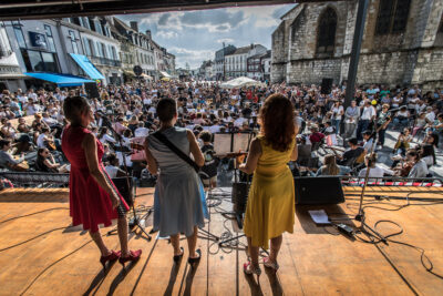 Trois femmes de dos debout sur une scène au premier plan, avec en fond la ville d'Orthez et un grand orchestre placé devant une foule de gens pour un concert. - Agrandir l'image 2 sur 6, fenêtre modale