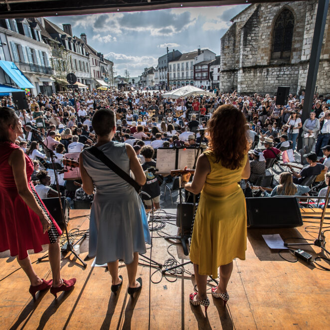 Trois femmes de dos debout sur une scène au premier plan, avec en fond la ville d'Orthez et un grand orchestre placé devant une foule de gens pour un concert.