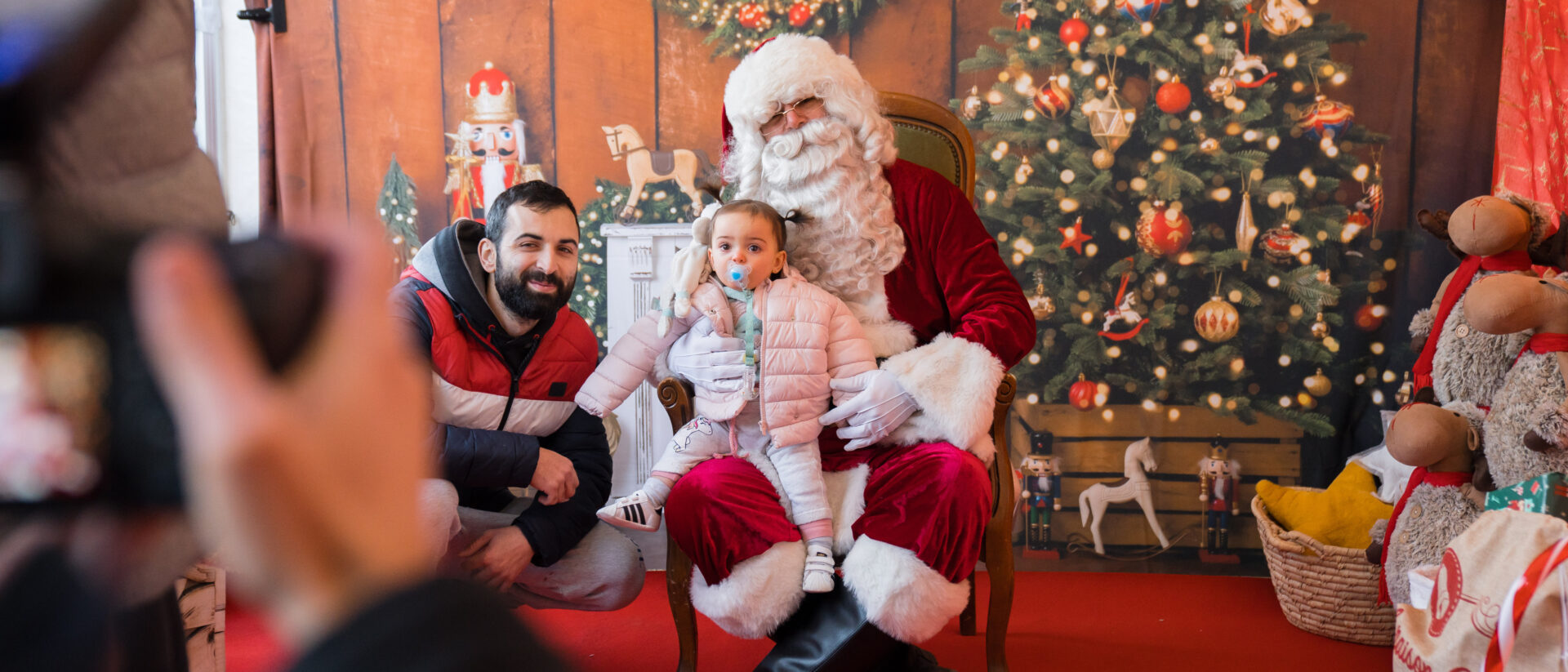 Séance photo avec sur la gauche de la photo, un bout de l'appreil photo et de la main du photographe, et face à nous un décor de Noël avec un sapin et des ton boisés comme dans un chalet, avec une petite fille en doudoune et une tétine dans la bouche assise sur les genoux du Père-Noël,, aux côtés de son père accroupi à côté d'elle, sur la gauche de l'image. Des peluches empilées viennent compléter le décor sur la droite - Agrandir l'image, fenêtre modale