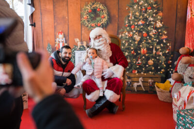 Séance photo devant un décor pour une petite assise sur les genoux du Père-Noël, aux côtés de son père. - Agrandir l'image 2 sur 3, fenêtre modale