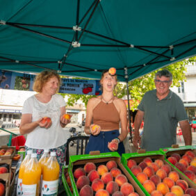 Trois producteurs locaux de fruits dans leur stand au marché, l’une d’entre-elles jongle avec des oranges.