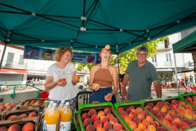 Trois producteurs locaux de fruit dans leur stand au marché, l’une d’entre-elles jongle avec des fruits. - Agrandir l'image 2 sur 6, fenêtre modale