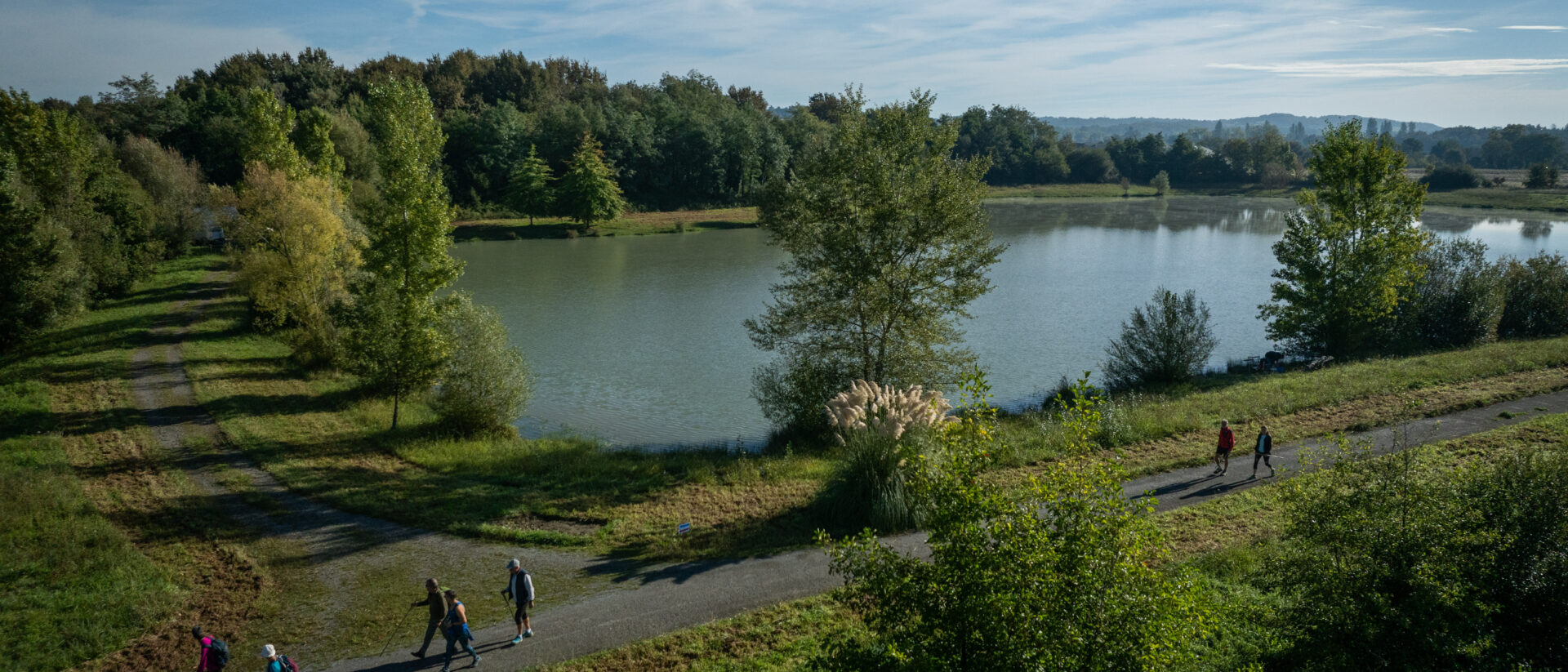 Randonneurs sur un chemin bétonné au bord d'un grand lac avec en arrière plan une forêt et un grand ciel bleu. - Agrandir l'image, fenêtre modale