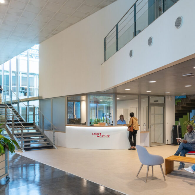 Personnes attendant dans la salle d'attente de l'accueil de la collectivité, en attendant qu'une dame termine d'échanger avec une agente d'accueil, pendant que deux agents sur la gauche dans l'escalier, échangent sur un dossier.