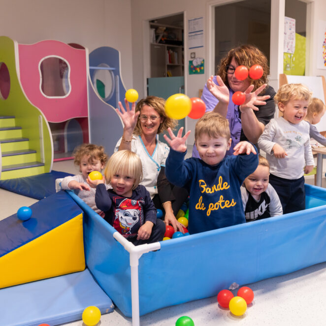 Cinq enfants et deux auxiliaires de puériculture dans une piscine à balle et les jetant partout dans la pièce.
