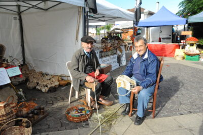Deux hommes âgés assis sur une chaise devant un stand, souriant à la caméra. - Agrandir l'image 2 sur 3, fenêtre modale