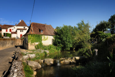 Vue d'une rue de la commune avec une vieille maison encore habitée avec des lières et un petit pont au dessus d'une rivière avec de gros rochers autour. - Agrandir l'image 1 sur 3, fenêtre modale