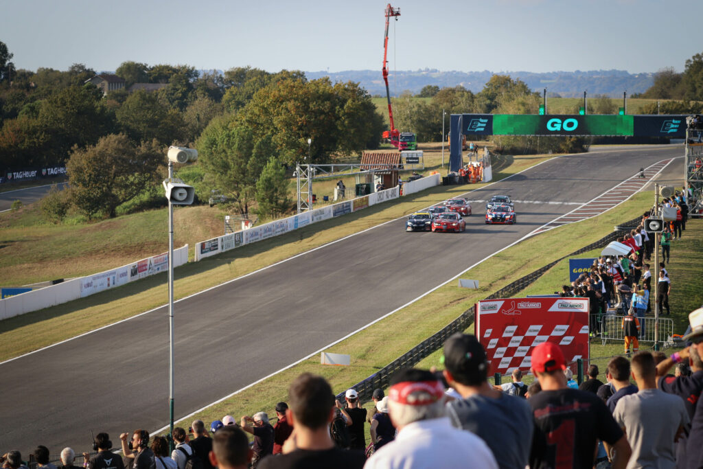 Vue depuis les gradins du démarrage d'une course automobile sur un circuit dédié avec en fond une belle campagne. - Agrandir l'image, fenêtre modale