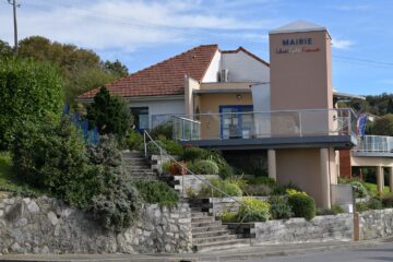 Vue sur le bâtiment de la mairie qui surplombe la route. Un escalier accessible depuis le trottoir permet de rejoindre l'entrée. Les valeurs de la république sont inscrite sur le haut d'un mur du bâtiment.