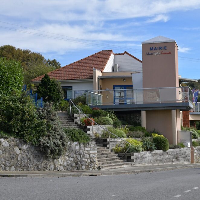 Vue sur le bâtiment de la mairie qui surplombe la route. Un escalier accessible depuis le trottoir permet de rejoindre l'entrée. Les valeurs de la république sont inscrite sur le haut d'un mur du bâtiment.