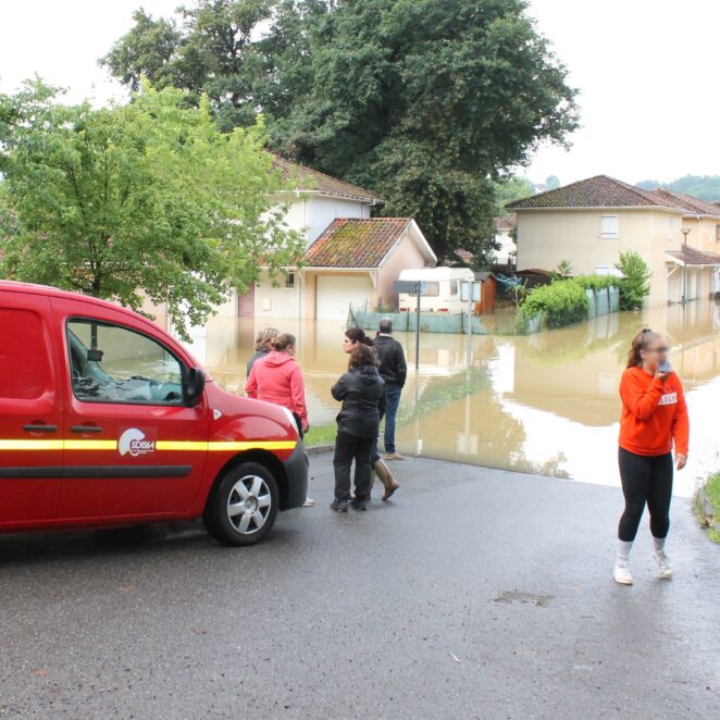 Groupe de personne attendant avec les sapeurs pompiers sur le bord d'une route inondée au milieu d'un petit quartier résidentiel.