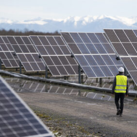 Homme habillé en tenue de travail avec un gilet jaune et un casque, marchant au milieu de grand panneaux photovoltaïques.