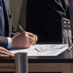 Homme signant un contrat sur table avec un trophée transparent devant lui.