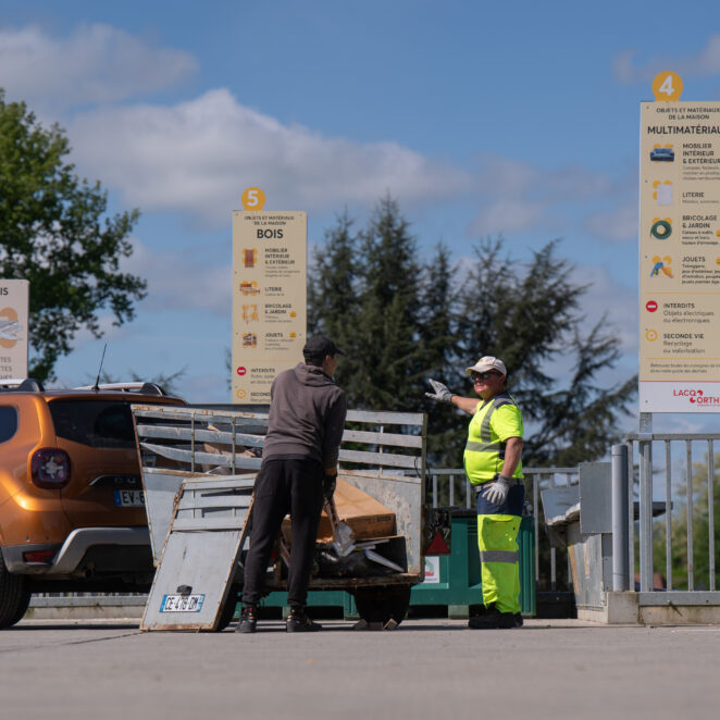 Un agent de déchetterie montrant du doigt à un habitant l'emplacement de la benne dans laquelle il doit jeter le contenu de sa remorque.