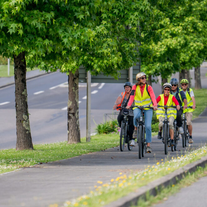 Groupe de cyclistes avec casque et gilets jaunes se promenant sur une piste cyclable sécurisée en plein cœur d'une commune et à côté d'une route.