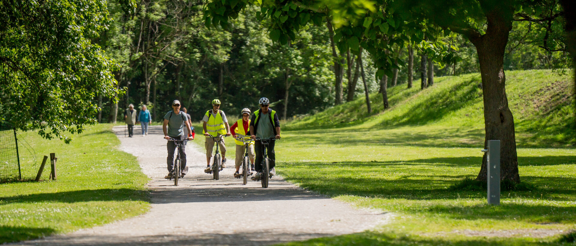 Groupe à vélo habillé en tenue correcte dont casque et gilet jaune vu de loin qui organise une balade sur un chemin dédié au milieu de la nature bien entretenue de la base de loisirs Orthez-Biron.