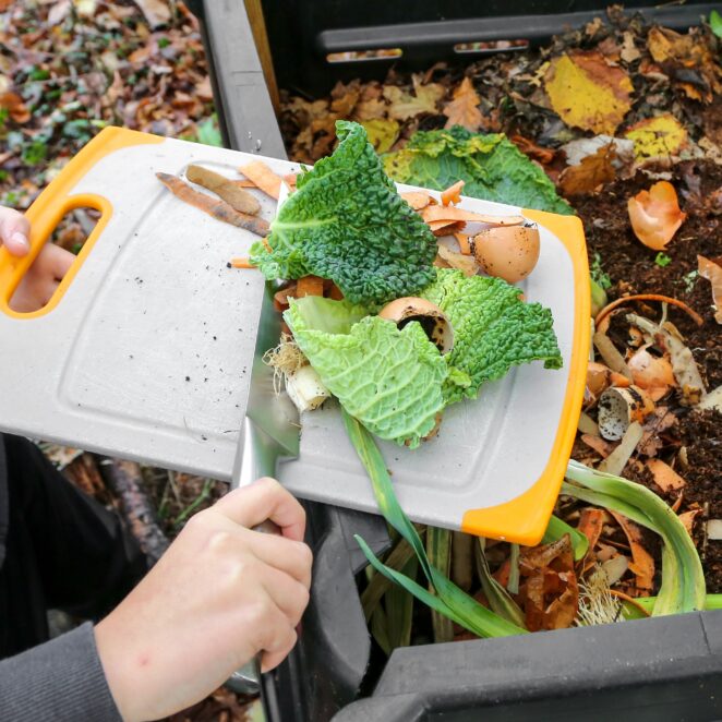 Personne faisant glisser à l'aide d'un couteau, des déchets alimentaires type salade et coquilles d'oeuf d'une planche à découper, dans un composteur en partie plein.