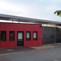 Rouge et grise, la médiathèque de Monein est un grand bâtiment de plein pied entouré d'un parking