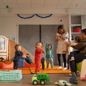 Dans une salle de crèche colorée et pleine de jeux, 3 enfants de moins de 3 ans se tiennent debout et s'amusent avec le personnel de crèche. Un autre enfant est assis sur les genou d'un deuxième agent.