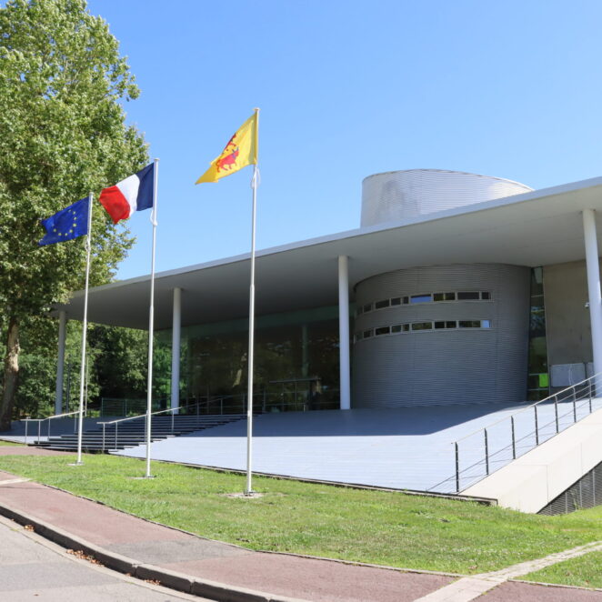 Photo de face du bâtiment de la communauté de communes à Mourenx. Une petite route passe juste devant le bâtiment. Des marches et une rampe permettent d'accéder à des portes vitrés