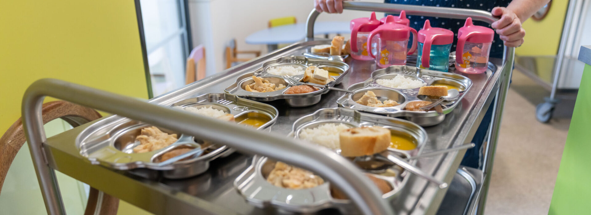 Un chariot de cantine en inox est poussé dans une salle. Il est rempli d’assiettes compartimentées en métal contenant des repas pour enfants (riz, légumes, soupe, pain…). On voit aussi plusieurs gobelets roses à bec avec de l’eau pour enfants. Une personne, visible seulement des épaules aux mains, pousse le chariot au sein d'une crèche. - Agrandir l'image, fenêtre modale