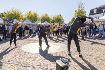 Trois performeurs du collectif StreetSmile s'échauffe le dos en se penchant sur la droite avec les bras en l'air, entourés par le public venu nombreux. - Agrandir l'image 5 sur 9, fenêtre modale
