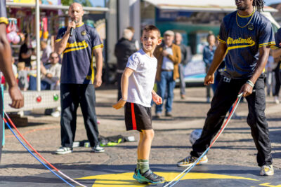 Un jeune enfant, qui sourit à la caméra, apprenant des chorégraphies basique du collectif avec une corde à sauté et aidé des artistes juste derrière lui. - Agrandir l'image 7 sur 9, fenêtre modale