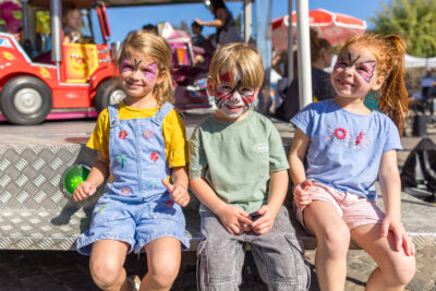 Trois enfants assis près d'un manège nous montrant fièrement leur maquillage artistique sur le visage. Les deux fillettes situées de part et d'autre de la photo ont un dessin de papillon et le petit garçon au centre a une araignée stylisée comme spiderman. - Agrandir l'image 9 sur 9, fenêtre modale