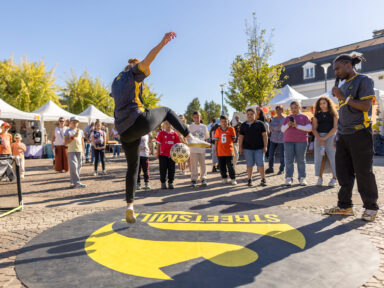 Un danseur du collectif StreetSmile de dos, entouré d'enfants qui regardent l'air émerveillé l'artiste qui jongle avec un ballon de foot et sa jambe droite. Un deuxième artiste du collectif est à sa droit et commente la scène au micro.