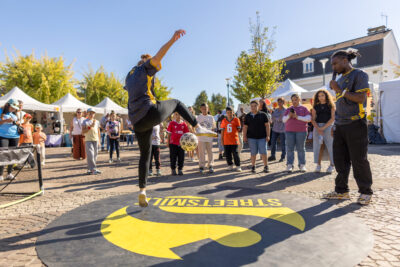 Un danseur du collectif StreetSmile de dos, entouré d'enfants qui regardent l'air émerveillé l'artiste qui jongle avec un ballon de foot et sa jambe droite. Un deuxième artiste du collectif est à sa droit et commente la scène au micro. - Agrandir l'image 6 sur 9, fenêtre modale