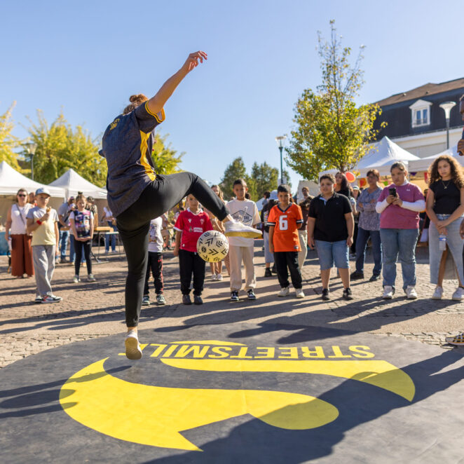 Un danseur du collectif StreetSmile de dos, entouré d'enfants qui regardent l'air émerveillé l'artiste qui jongle avec un ballon de foot et sa jambe droite. Un deuxième artiste du collectif est à sa droit et commente la scène au micro.