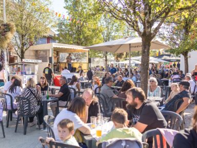 Plusieurs tables en plastique toutes occupées par des personnes en terrasse qui profitent d'un petit concert sur scène à l'ombre des arbres avec un beau ciel bleu.