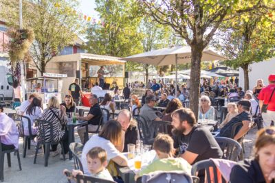 Plusieurs tables en plastique toutes occupées par des personnes en terrasse qui profitent d'un petit concert sur scène à l'ombre des arbres avec un beau ciel bleu. - Agrandir l'image 1 sur 9, fenêtre modale