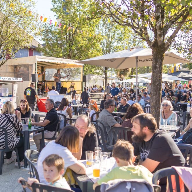Plusieurs tables en plastique toutes occupées par des personnes en terrasse qui profitent d'un petit concert sur scène à l'ombre des arbres avec un beau ciel bleu.