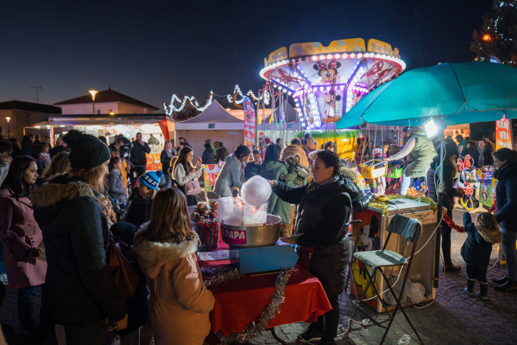 Vue de nuit du marché de Noël d'Artix avec une femme préparant une barbe à papa et derrière elle, un manège pour enfant. - Agrandir l'image, fenêtre modale