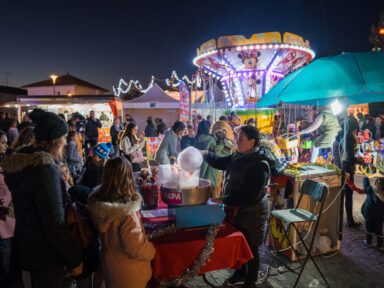 Vue de nuit du marché de Noël d'Artix avec une femme préparant une barbe à papa et derrière elle, un manège pour enfant.