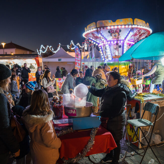 Vue de nuit du marché de Noël d'Artix avec une femme préparant une barbe à papa et derrière elle, un manège pour enfant.