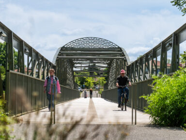 Vue de la passerelle de face, prise de vue du sol où l'on voit droit devant nous le chemin sur le pont, toute l'architecture de la passerelle qui forme une arche de métal en dessous de laquelle se trouve au loin deux piétons, et devant nous relativement proches, une femme qui marche sur la gauche de l'image, et un homme à vélo sur la droite, en train de se diriger vers nous. Tout autour de la passerelle on distingue de la verdure et des buissons.
