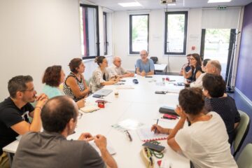 Petite salle avec une table ovale au milieu avec tout autour des femmes et des hommes qui échangent sur un sujet avec devant eux sur la table, de quoi prendre des notes donc des feuilles et stylos.