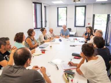 Petite salle avec une table ovale au milieu avec tout autour des femmes et des hommes qui échangent sur un sujet avec devant eux sur la table, de quoi prendre des notes donc des feuilles et stylos.