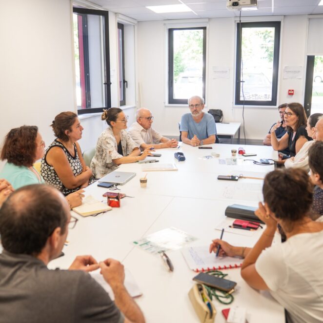 Petite salle avec une table ovale au milieu avec tout autour des femmes et des hommes qui échangent sur un sujet avec devant eux sur la table, de quoi prendre des notes donc des feuilles et stylos.