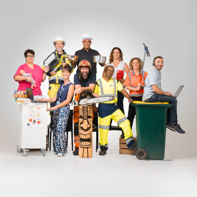 9 agents de la collectivité issus de métiers différents, posent en tenue de travail sur un fond uni. De gauche à droite, on voit une assistante maternelle avec un poupon dans la main gauche et un biberon dans la main droite, à côté un agent des services techniques avec des machines dans les mains qui ressemble à une tablette, à côté un cuisinier avec sa toque et deux récipient un dans chaque main, à côté une femme des services administratifs en tenue simple mais tenant en main le code de l'urbanisme, à côté une femme des services techniques avec une pioche dans les mains, à côté un agent d'accueil avec un micro-casque et un ordinateur sur les genoux, il est assis sur une poubelle au couvercle jaune. A gauche de la poubelle se tient un agent du service déchets tenant la poubelle, à sa gauche un agent des services techniques accoudé sur un totem de bois et tenant une tronçonneuse, et à sa gauche, une médiathécaire habillée décontracté avec un ordinateur devant elle et un support roulant avec une affiche du centre culturel Le MIX accroché dessus et transportant divers bouquins.