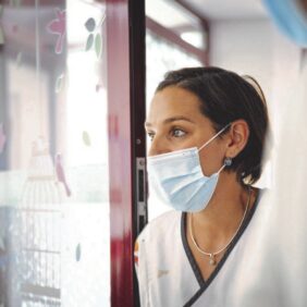 Femme issu du corps médical en blouse blanche et masque chirurgical sur la droite de la photo, regardant vers la gauche à travers une fenêtre.