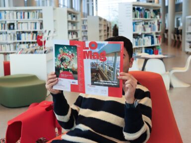 Jeune homme assis jambes croisées dans un fauteuil au milieu d'une grande médiathèques avec des bibliothèques de part et d'autres de la photo, il lit le dernier magazine de Lacq-Orthez en cachant son visage.