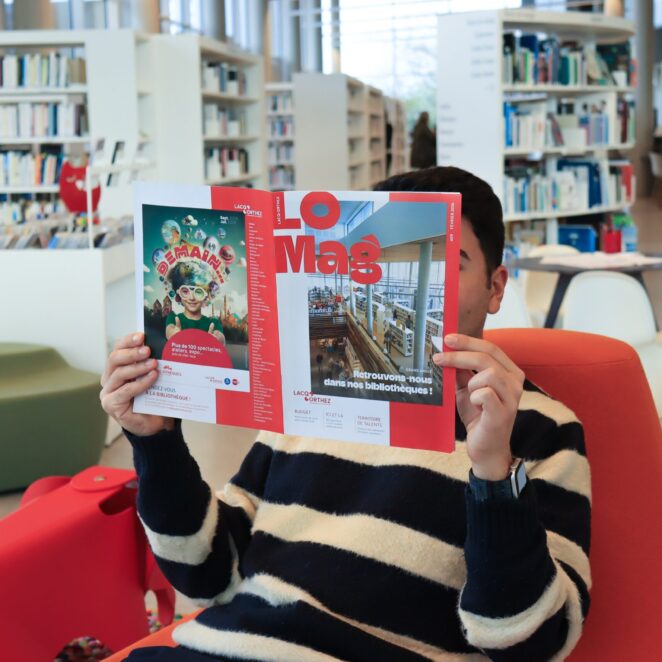 Jeune homme assis jambes croisées dans un fauteuil au milieu d'une grande médiathèques avec des bibliothèques de part et d'autres de la photo, il lit le dernier magazine de Lacq-Orthez en cachant son visage.