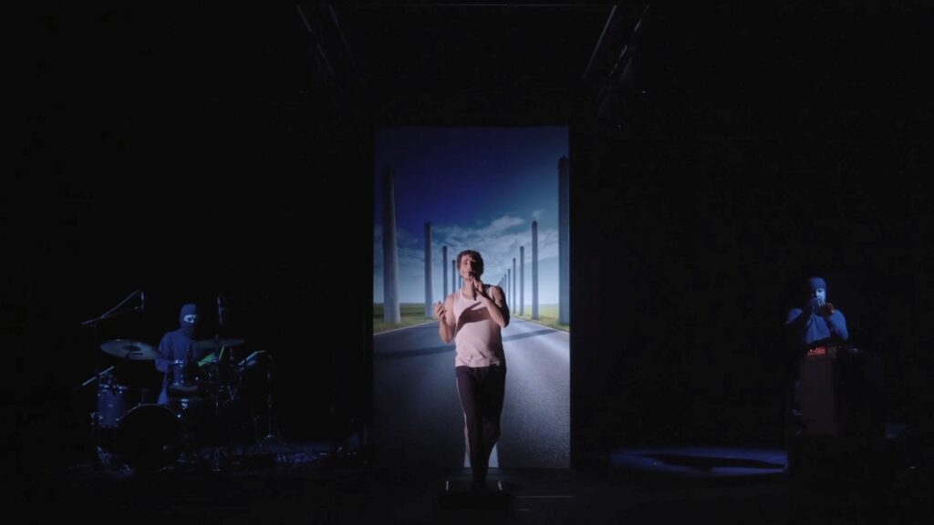 Scène de spectacle dans le noir, à gauche éclairé légèrement, le batteur, à droite éclairé légèrement, le trompettiste, et au centre un jeune homme qui chante au micro, éclairé en lumière blanche avec derrière lui une projection à la verticale, d'un décor de route qui accompagne ses paroles et va changer au fur et à mesure du spectacle pour s'immerger dans l'univers.