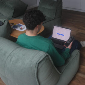 Jeune homme assis dans un fauteuil de salon avec son ordinateur portable sur les genoux et son téléphone en main