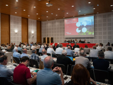 Salle de réunion remplie avec face à nous les élus, de dos, assis en rangées, se tenant face au bureau principal et à un diaporama qui défile grâce à un projecteur.