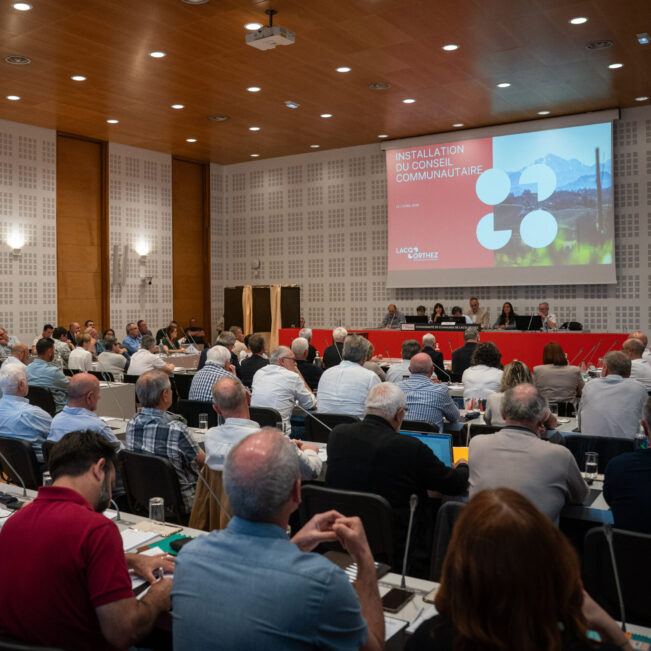 Salle de réunion remplie avec face à nous les élus, de dos, assis en rangées, se tenant face au bureau principal et à un diaporama qui défile grâce à un projecteur.
